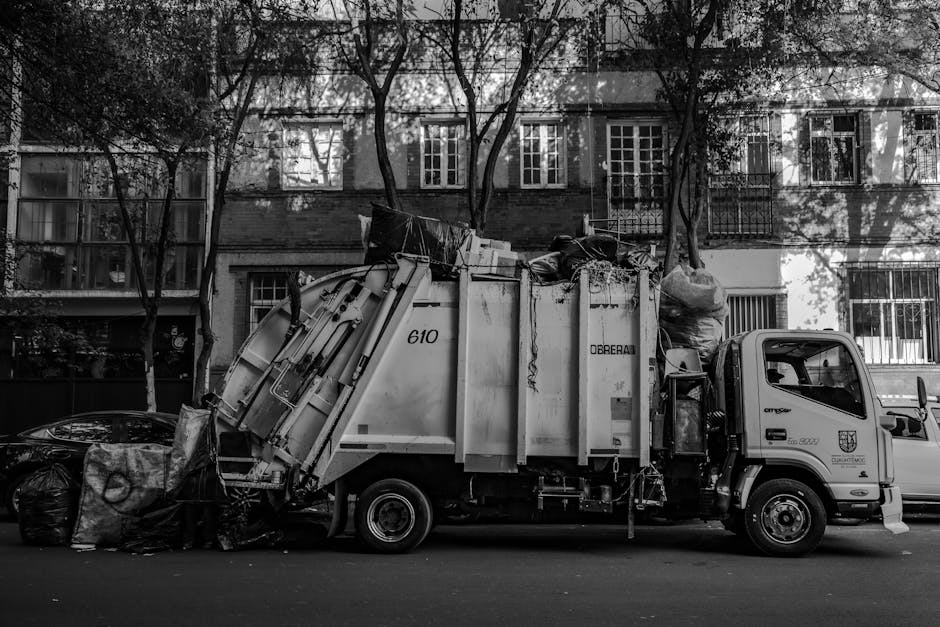 A worker in a red and yellow high-visibility jacket is standing beside a large red rubbish collection truck, which is parked at the side of a paved road. The back of the truck is open, revealing mecha